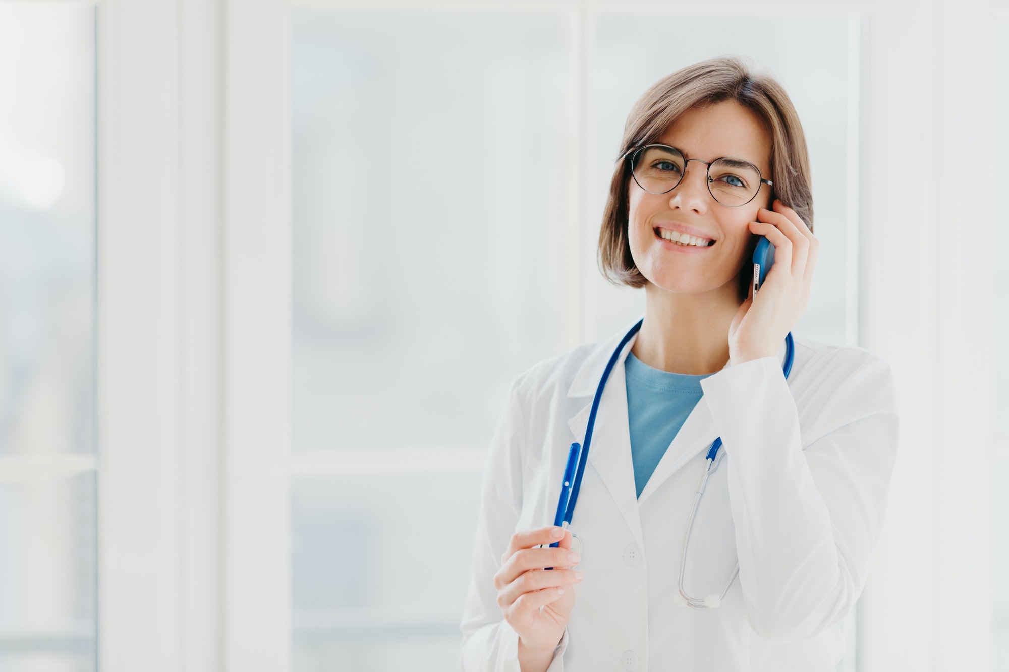 Positive woman doctor consults patient via cellphone, looks gladfully